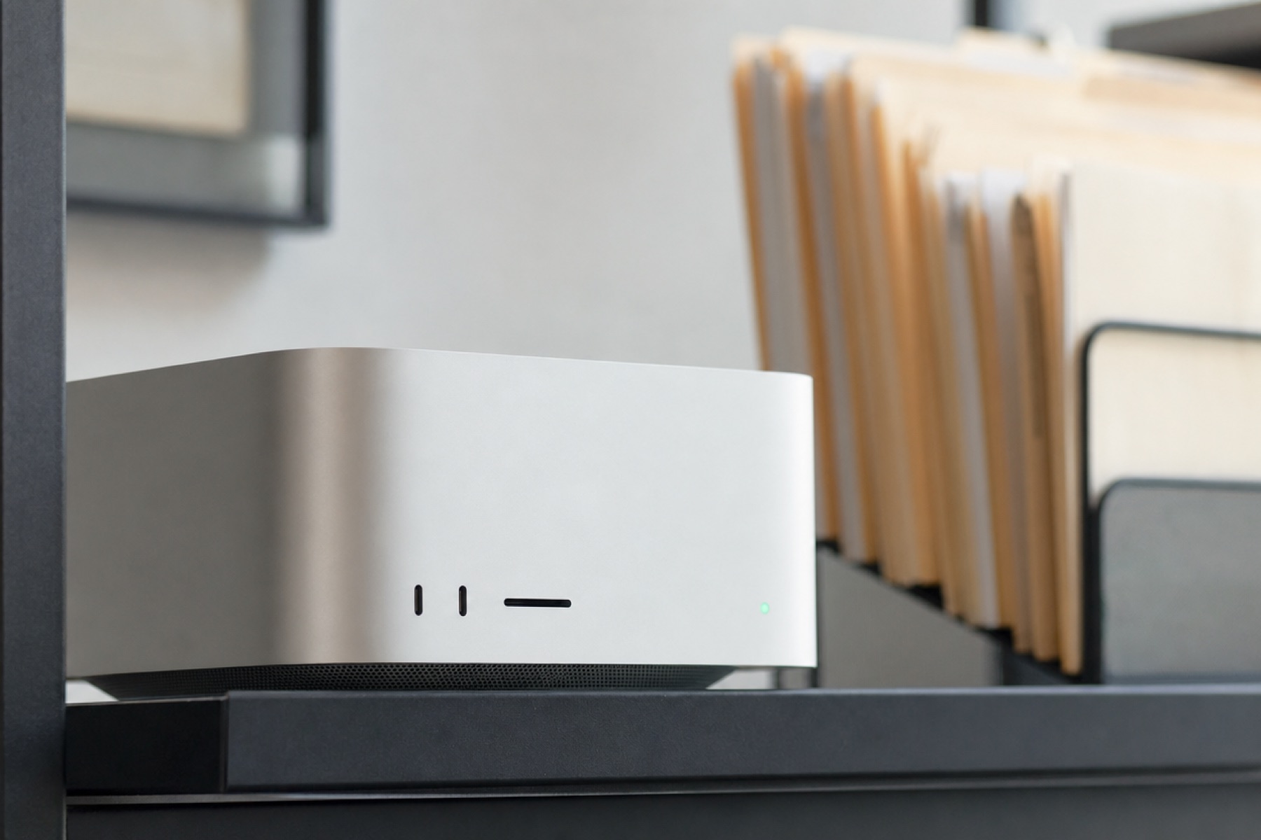 A Mac Studio on an office shelf, its status light on, paper folders behind it.